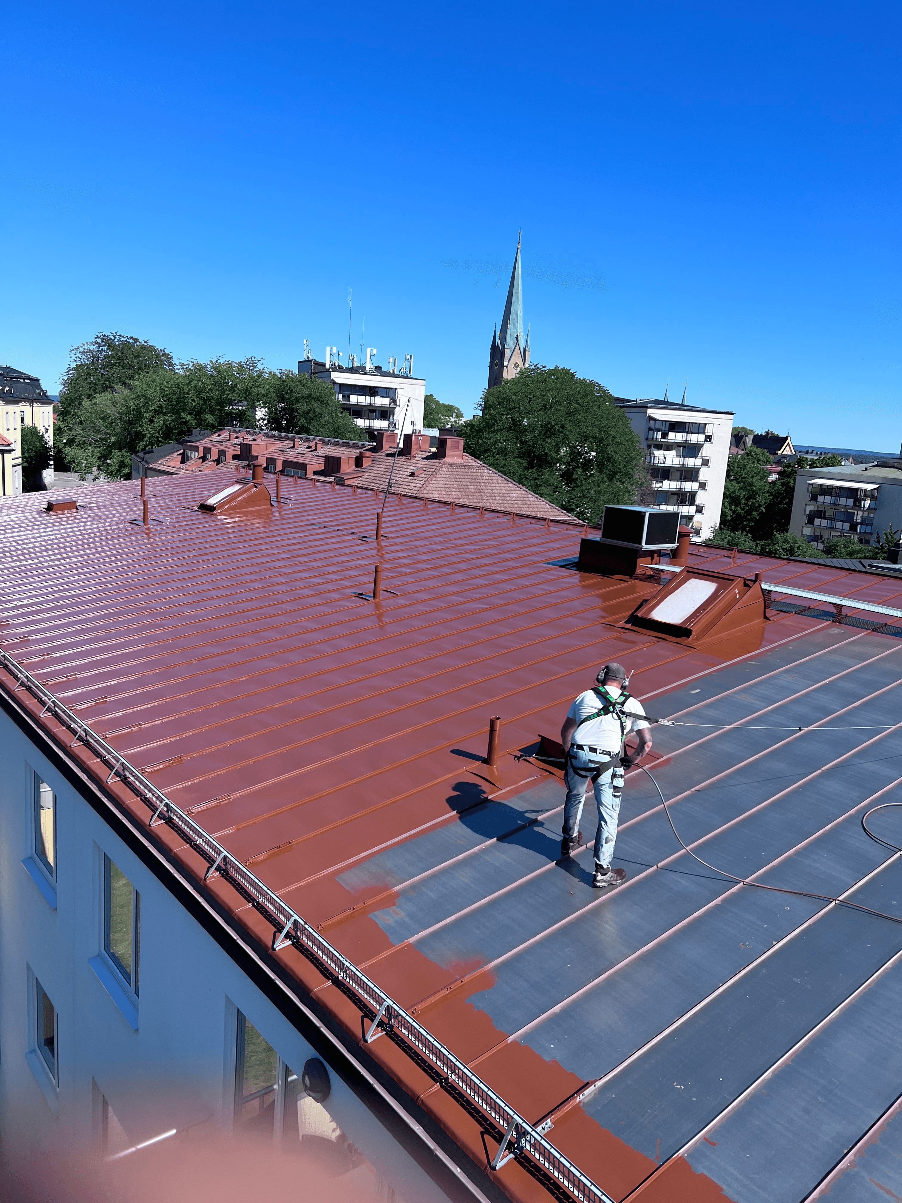 Hantverkare i sele arbetar på ett stort rostrött plåttak, med Linköpings domkyrka i bakgrunden